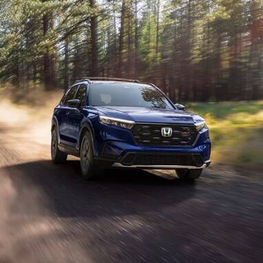2027 Honda CR-V TrailSport Hybrid in Canyon River Blue Metallic front driving through a sunlit forest on a dirt road (Compact Crossover SUV).