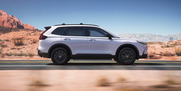 2027 Honda CR-V Sport Touring Hybrid in Platinum White Pearl profile driving along a scenic road (Compact Crossover SUV).